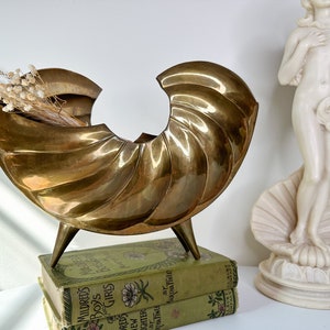 May include: A gold-colored brass seashell-shaped decorative bowl with a textured surface. The bowl is on a stack of two books with floral designs. A white porcelain statue of a woman is in the background.