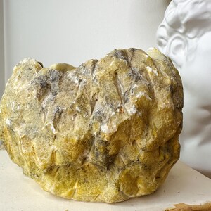 May include: A large, irregularly shaped, yellow-green stone bowl with a rough, textured surface. The bowl has areas of gray and white, suggesting natural mineral deposits. The bowl is sitting on a stack of books.