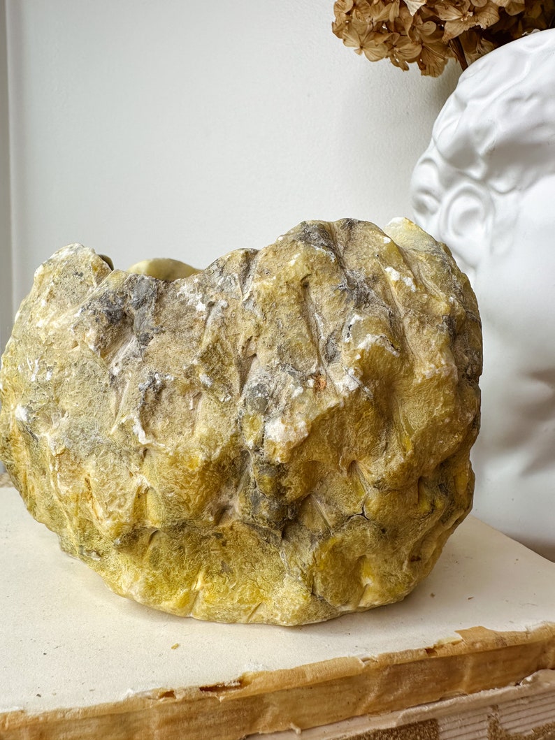 May include: A large, irregularly shaped, yellow-green stone bowl with a rough, textured surface. The bowl has areas of gray and white, suggesting natural mineral deposits. The bowl is sitting on a stack of books.