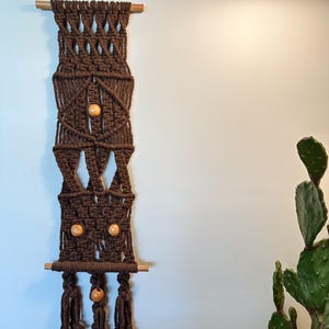 May include: A dark brown macrame wall hanging featuring intricate knotted patterns, suspended from a horizontal wooden dowel. The design incorporates four smooth, light-colored wooden beads integrated into the fiber structure, ending in three tassels.