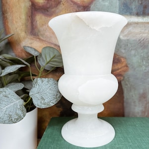 May include: A white alabaster urn vase featuring a wide, flared rim, a rounded central body, and a pedestal base. The stone has a smooth, polished finish with subtle, natural translucent veining throughout the material, creating a soft glow.