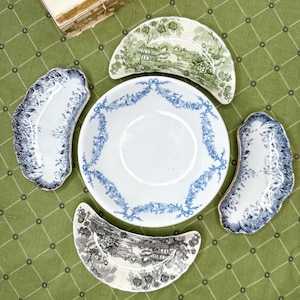 May include: A collection of antique porcelain dishes. The set includes a large white plate with blue floral detailing, and four smaller, uniquely shaped plates with landscape and floral designs in blue, green, and black. The dishes are arranged on a green tablecloth.