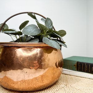 May include: A large, antique copper cauldron with a curved handle, filled with faux green foliage. The cauldron has a shiny, reflective surface and sits on a woven mat. A green book is visible in the background.
