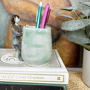 May include: A light green ceramic pencil holder featuring a small, attached figurine of a person in a kneeling pose. The holder contains one teal pen and one pink pen, and it is positioned on top of a white book with gold lettering.