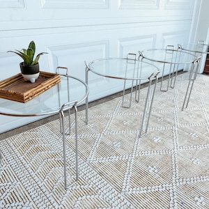 May include: Set of four nesting tables with clear glass tops and chrome legs. The tables have a round design and are arranged in a row. One table has a decorative tray with a plant on top. The tables are 20 inches in diameter.