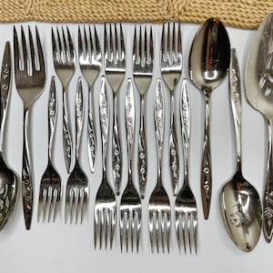 May include: A collection of stainless steel flatware including various forks, spoons, and a slotted serving utensil. Each piece features a polished metallic finish and a delicate floral pattern embossed along the handle stems.