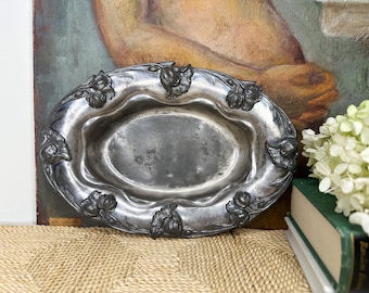 May include: An oval-shaped silver-toned metal bowl featuring a raised floral relief pattern along the wide, undulating rim. The center of the bowl is smooth and slightly recessed, while the exterior border is decorated with multiple flower motifs.