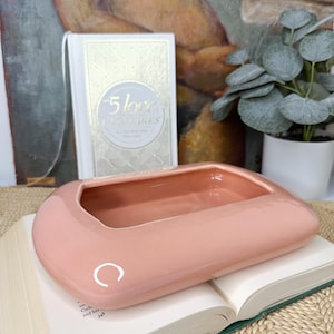 May include: A pale pink ceramic dish with a rounded rectangular shape and a slightly raised rim. The dish has a smooth, glossy finish and a subtle curve in the center. The dish is sitting on top of a closed book.