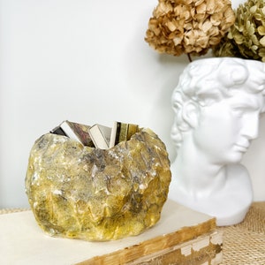 May include: A yellow and gray stone-like bowl filled with small books. The bowl sits on top of a stack of old books. A white bust and dried flowers are in the background.