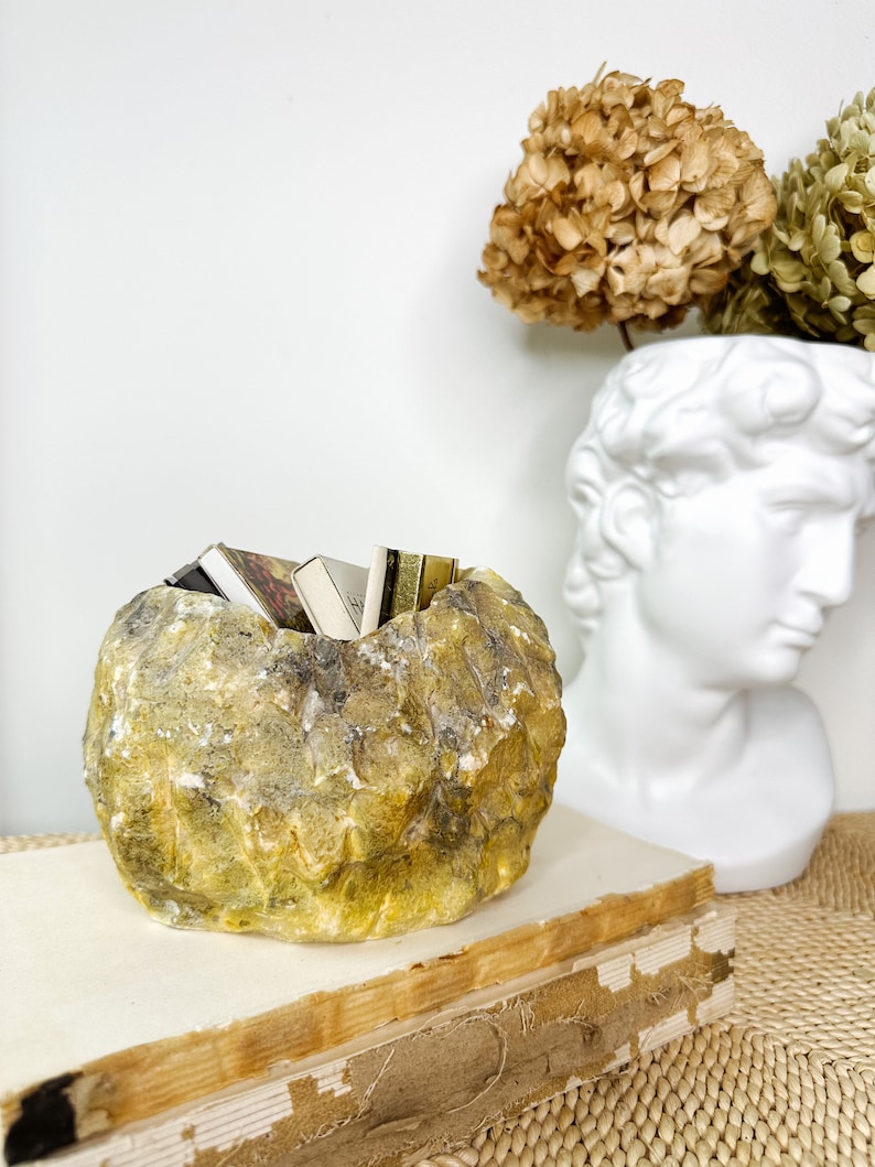May include: A yellow and gray stone-like bowl filled with small books. The bowl sits on top of a stack of old books. A white bust and dried flowers are in the background.