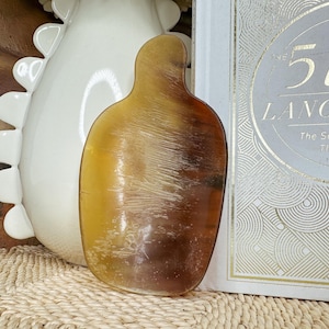 May include: A translucent, amber-colored, organic-shaped object with a smooth surface. The object has a gradient of colors, transitioning from a light yellow to a deep brown. It is next to a white decorative vase and a book titled "The 5 Love Languages".