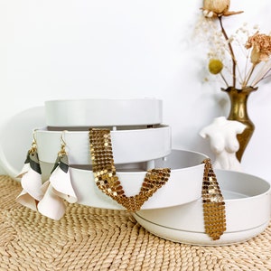 May include: A white tiered jewelry organizer with three circular trays. A pair of layered earrings with white, gray, and tan petals hangs from the side. A gold-toned metallic mesh chain is draped over the edges of the middle and bottom trays.