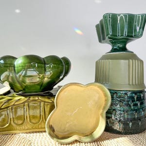 May include: A collection of vintage ceramic decorative items in shades of green and yellow. The items include a scalloped-edge dish, a pedestal vase, and two decorative bowls. The items are arranged on a woven surface.