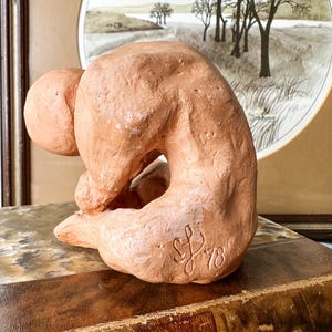 May include: A terracotta sculpture of a seated figure in a curled position. The sculpture has a rough texture and is signed "SL '78". The figure is placed on a vintage book with gold lettering, with a framed landscape painting in the background.