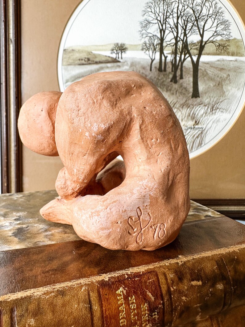 May include: A terracotta sculpture of a seated figure in a curled position. The sculpture has a rough texture and is signed "SL '78". The figure is placed on a vintage book with gold lettering, with a framed landscape painting in the background.