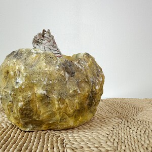 May include: A natural, rough-hewn stone bowl in shades of yellow and brown, with a bundle of white sage tied with red string resting inside. The bowl sits on a woven mat, creating a rustic, earthy aesthetic.