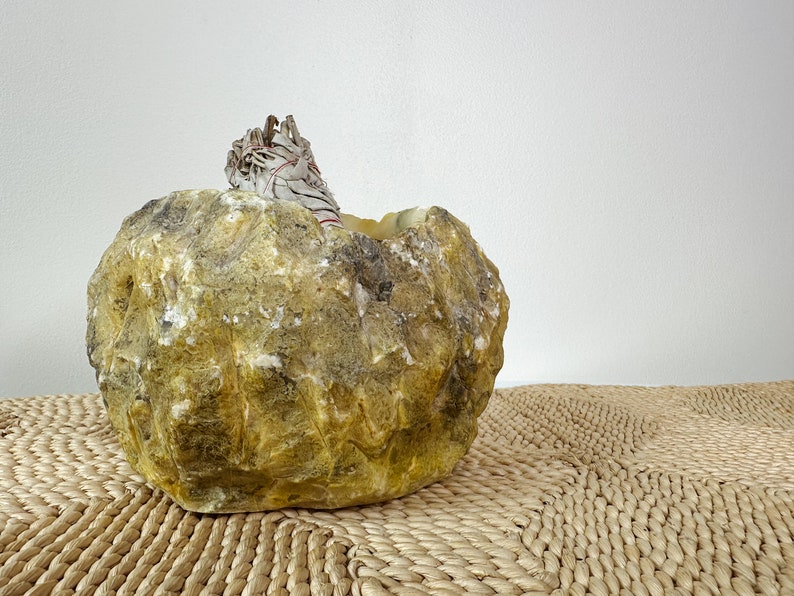 May include: A natural, rough-hewn stone bowl in shades of yellow and brown, with a bundle of white sage tied with red string resting inside. The bowl sits on a woven mat, creating a rustic, earthy aesthetic.