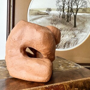 May include: A terracotta-colored abstract sculpture with a large central opening. The sculpture has a rough, textured surface and sits on a vintage book. A framed landscape painting with trees and a river is in the background.