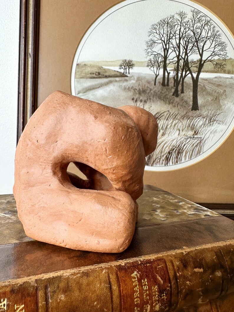 May include: A terracotta-colored abstract sculpture with a large central opening. The sculpture has a rough, textured surface and sits on a vintage book. A framed landscape painting with trees and a river is in the background.
