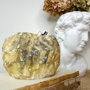 May include: A textured, yellow-toned stone sculpture with dark gray and white accents. The sculpture is rectangular with carved lines. A white bust of a classical figure and dried flowers are in the background.