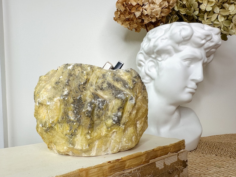 May include: A textured, yellow-toned stone sculpture with dark gray and white accents. The sculpture is rectangular with carved lines. A white bust of a classical figure and dried flowers are in the background.