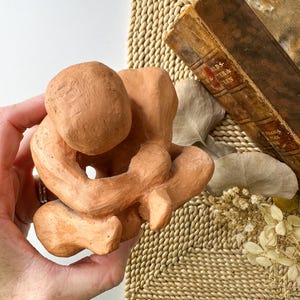 May include: A terracotta-colored sculpture of a person in a seated, curled position. The figure is holding its knees, with the head resting on the knees. The sculpture has a rough, textured surface and is held in a hand.