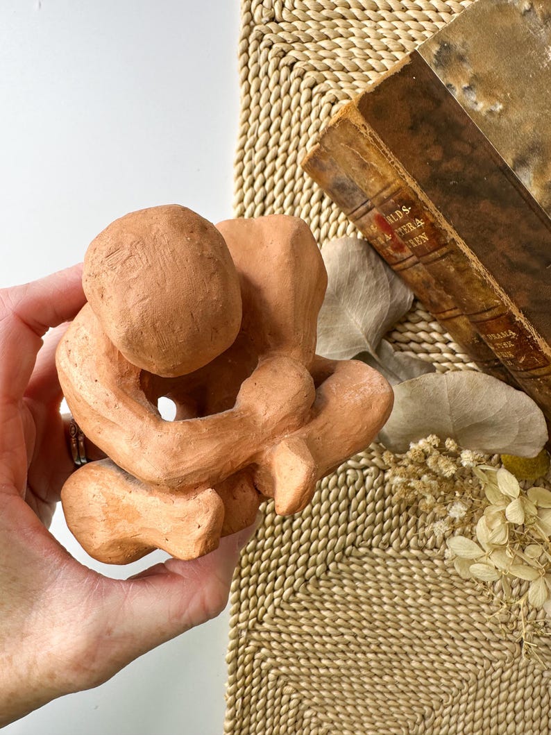 May include: A terracotta-colored sculpture of a person in a seated, curled position. The figure is holding its knees, with the head resting on the knees. The sculpture has a rough, textured surface and is held in a hand.