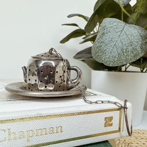 May include: A silver-toned metal tea infuser shaped like a miniature teapot with a matching saucer. The infuser features a perforated body and lid, a small handle, and a spout, connected by a thin metal chain to a circular ring for easy handling.