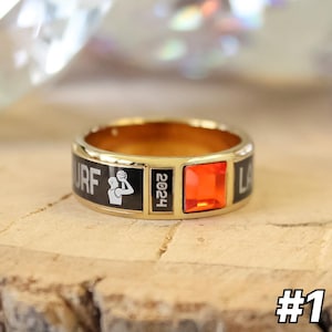 May include: A gold-colored ring with a black band featuring white text and a basketball player graphic. The ring has a square, orange gemstone and the year "2024" is displayed. The ring is set on a wooden surface.