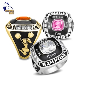 May include: Three different championship rings with black enamel and white stones. The first ring features a gold band with a cheerleader design. The second ring has a pink stone and the text "Your Message Here". The third ring has a clear stone and the text "Kevin 2024".