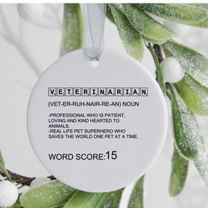 May include: White round ornament with black text that reads "VET, ERI, NA, RIA, N (VET-ER-RUH-NAIR-RE-AN) NOUN -PROFESSIONAL WHO IS PATIENT, LOVING AND KIND HEARTED TO ANIMALS. -REAL LIFE PET SUPERHERO WHO SAVES THE WORLD ONE PET AT A TIME. WORD SCORE:15"