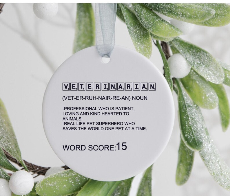 May include: White round ornament with black text that reads "VET, ERI, NA, RIA, N (VET-ER-RUH-NAIR-RE-AN) NOUN -PROFESSIONAL WHO IS PATIENT, LOVING AND KIND HEARTED TO ANIMALS. -REAL LIFE PET SUPERHERO WHO SAVES THE WORLD ONE PET AT A TIME. WORD SCORE:15"