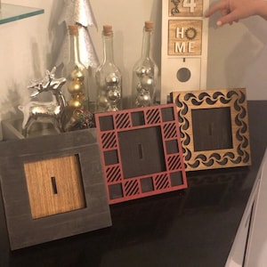 May include: Three wooden picture frames with different designs. The frames are black, red, and brown. The frames are on a black surface. The text "Party of 4" and "Home" is visible on a white sign in the background.