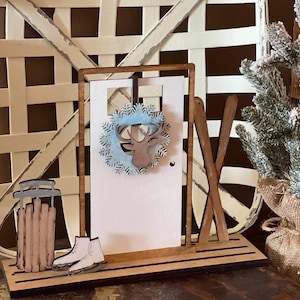 May include: Wooden Christmas decor with a white door, a wreath with a reindeer, skis, a sled, and two ice skates. The decor is set on a wooden base.