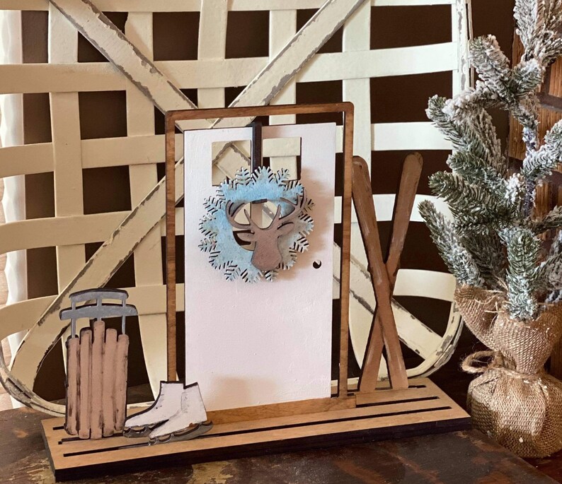 May include: Wooden Christmas decor with a white door, a wreath with a reindeer, skis, a sled, and two ice skates. The decor is set on a wooden base.