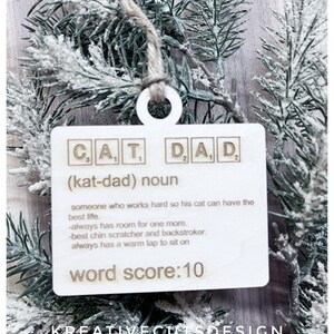 May include: A white wooden ornament with the word "CAT DAD" spelled out in Scrabble tiles. The ornament also includes the definition of a "cat dad" and a word score of 10.