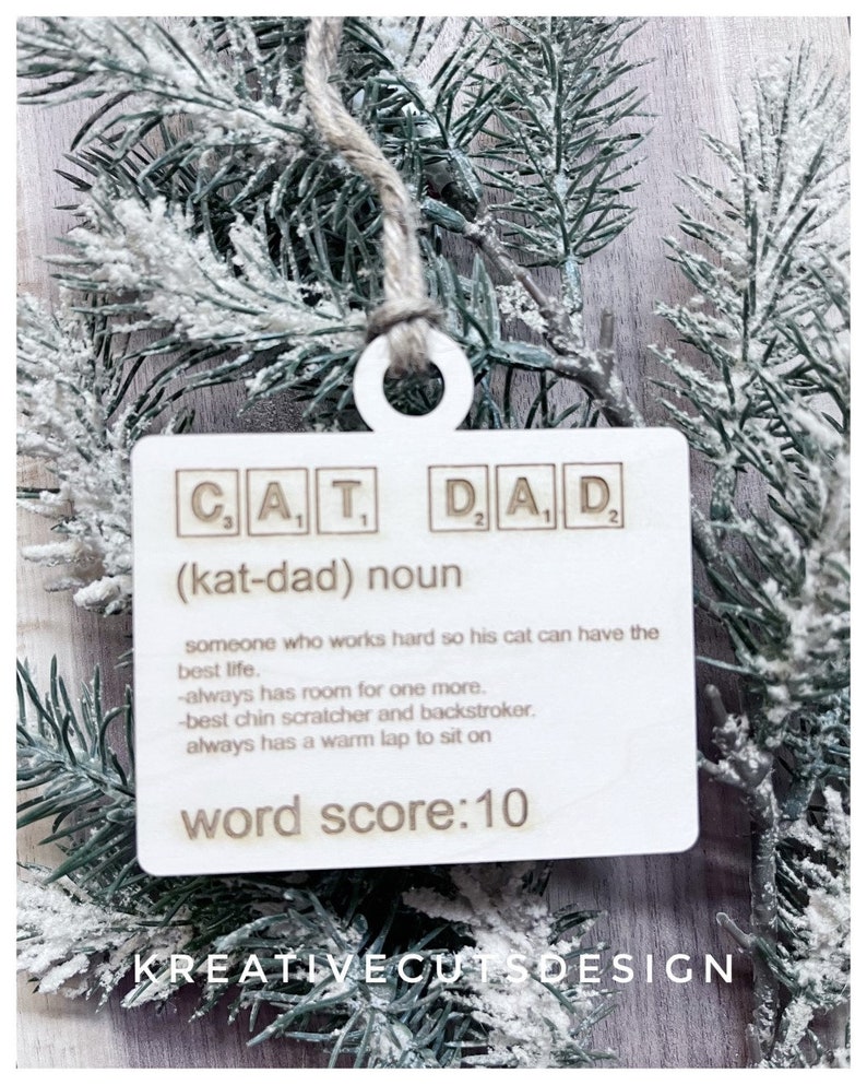 May include: A white wooden ornament with the word "CAT DAD" spelled out in Scrabble tiles. The ornament also includes the definition of a "cat dad" and a word score of 10.