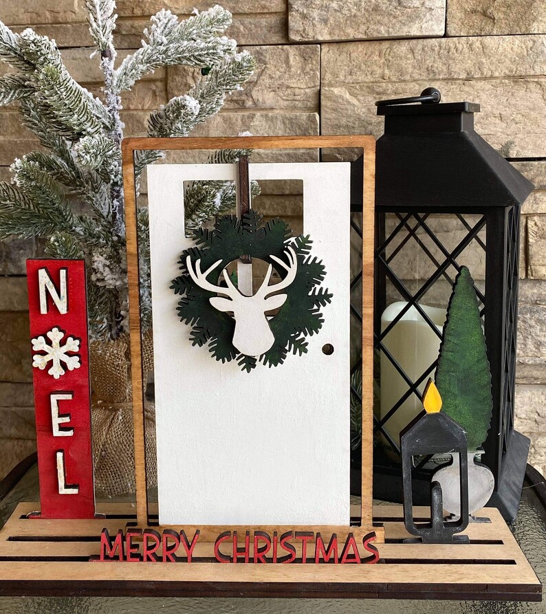 May include: Wooden Christmas decor with a white door, a green wreath with a deer silhouette, a red "NOEL" sign, and a black lantern with a candle.