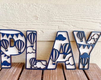 May include: Wooden letters spelling "PLAY" with a hot air balloon theme. The letters are navy blue with white interiors, featuring hot air balloons, clouds, and bunting. A decorative piece for a nursery or playroom.