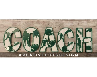 Peut inclure: Lettres en bois vertes et blanches épellant "COACH" avec une silhouette de joueurs de football américain et d'équipements à l'intérieur de chaque lettre. Les lettres sont sur un fond en bois avec le texte "KREATIVECUTSDESIGN" en dessous.