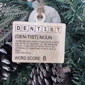 May include: A wooden Christmas ornament shaped like a Scrabble tile with the word "DENTIST" spelled out. The ornament also includes a definition of the word, "(DEN-TIST) NOUN - SOMEONE WHO DOES THINGS WITH A SMILE AND GETS TO THE ROOT OF THE PROBLEM. -EQUALLY SKILLED AT FIXING TEETH, AS LISTENING TO ANNOYING PEOPLE WITH A SMILE. -DRILLING NINJA." and a word score of 8.