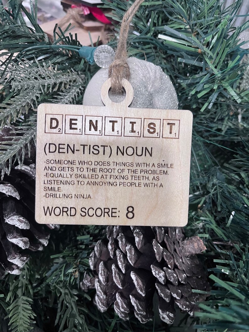 May include: A wooden Christmas ornament shaped like a Scrabble tile with the word "DENTIST" spelled out. The ornament also includes a definition of the word, "(DEN-TIST) NOUN - SOMEONE WHO DOES THINGS WITH A SMILE AND GETS TO THE ROOT OF THE PROBLEM. -EQUALLY SKILLED AT FIXING TEETH, AS LISTENING TO ANNOYING PEOPLE WITH A SMILE. -DRILLING NINJA." and a word score of 8.