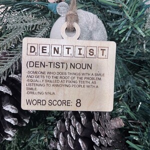 May include: A wooden Christmas ornament shaped like a Scrabble tile with the word "DENTIST" spelled out. The ornament also includes a definition of the word, "(DEN-TIST) NOUN - SOMEONE WHO DOES THINGS WITH A SMILE AND GETS TO THE ROOT OF THE PROBLEM. -EQUALLY SKILLED AT FIXING TEETH, AS LISTENING TO ANNOYING PEOPLE WITH A SMILE. -DRILLING NINJA." and a word score of 8.