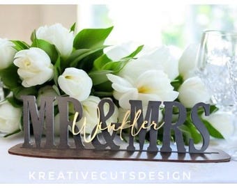 May include: A black wooden sign with the words "Mr. & Mrs. Winkler" in gold lettering. The sign is sitting on a wooden base and is surrounded by white tulips.