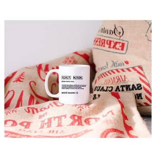 May include: White ceramic mug with a black and white design that says "CAT MOM" and defines the word as "(of a human) a person who loves cats" with a word score of 12. The mug is sitting on a burlap fabric with red and white Christmas-themed designs.