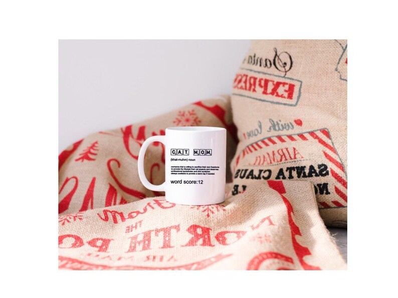 May include: White ceramic mug with a black and white design that says "CAT MOM" and defines the word as "(of a human) a person who loves cats" with a word score of 12. The mug is sitting on a burlap fabric with red and white Christmas-themed designs.