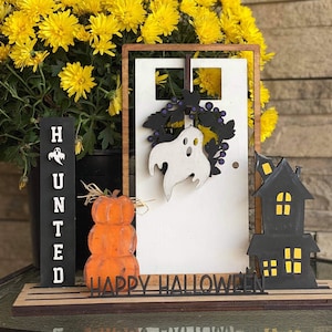 May include: A wooden Halloween sign with a white door, a ghost, and a black house. The sign says "Happy Halloween" and has a stack of pumpkins in front of it.