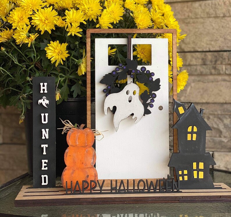 May include: A wooden Halloween sign with a white door, a ghost, and a black house. The sign says "Happy Halloween" and has a stack of pumpkins in front of it.