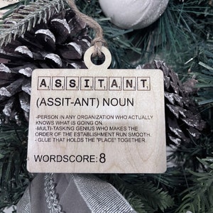 May include: Wooden Christmas ornament with the word "ASSISTANT" spelled out in Scrabble tiles. The ornament also includes the definition of the word, "Person in any organization who actually knows what is going on. Multi-tasking genius who makes the order of the establishment run smooth. Glue that holds the "place" together." and the wordscore, "WORDSCORE: 8".