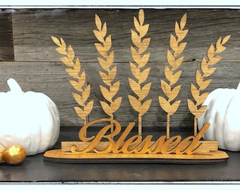 May include: Wooden sign with the word "Blessed" in a cursive font, surrounded by wheat stalks. The sign is sitting on a table with three white pumpkins and two orange pumpkins.